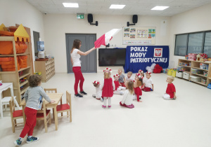 Dzieci siedzą na podłodze. Pani stoi, pokazuje dzieciom flagę Polski.